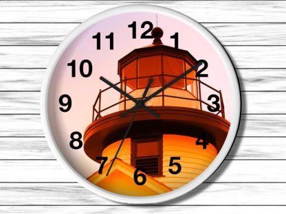 A round nautical wall clock with black numbers and hands features an image of a lighthouse at sunset. The warm orange glow creates a serene, peaceful ambiance.