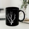 A black coffee mug featuring a white silhouette of a bare tree, set against a softly blurred background with plant.
