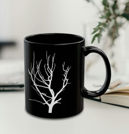 A black coffee mug featuring a white silhouette of a bare tree, set against a softly blurred background with plant.