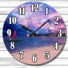 A clock with black numbers and hands features a serene seaside scene. The background has a pink and purple sky, birds flying, a lighthouse, and a boat with a red sail.
