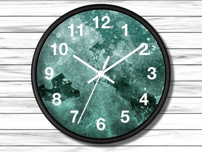 A round wall clock with a black rim features white numerals and hands against a teal green watercolor background. The clock is mounted on a white wooden wall.