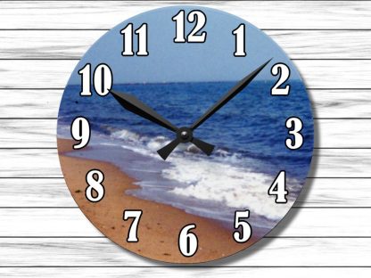 Round clock with beach scene featuring sandy shore, gentle waves, and blue ocean. Black hands and bold numbers on a wooden background. Peaceful vibe.