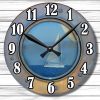 Rusted porthole clock with bold black numbers and hands. The face shows a serene sailboat on a blue sea, evoking nautical adventure. White wood background.