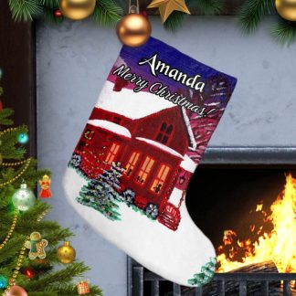 Personalized Christmas stocking with "Merry Christmas" text hangs by a lit fireplace. A painted house in a winter landscape adorns the stocking. A decorated tree with ornaments is on the left, creating a festive, cozy atmosphere.