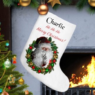 A festive Christmas stocking with Santa within a wreath and personalized text hangs by a roaring fireplace. A decorated Christmas tree with colorful lights and ornaments is nearby. Cozy and cheerful.