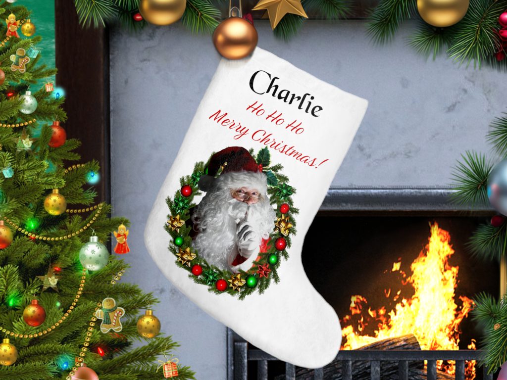 A festive Christmas stocking with Santa within a wreath and personalized text hangs by a roaring fireplace. A decorated Christmas tree with colorful lights and ornaments is nearby. Cozy and cheerful.