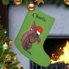 A personalized green Christmas stocking labeled "Charlie" hangs by a fireplace. It features a squirrel in a Santa hat. Nearby, a decorated Christmas tree glows warmly.