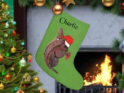 A personalized green Christmas stocking labeled "Charlie" hangs by a fireplace. It features a squirrel in a Santa hat. Nearby, a decorated Christmas tree glows warmly.