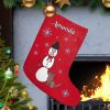 Personalized Red Christmas stocking with a snowman and a dog design hangs by a lit fireplace. Nearby, a decorated tree sparkles with colorful lights and ornaments. Cozy and festive atmosphere.