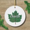 White, round ornament featuring a green leaf with "Charlie" in white script. Surrounded by pine branches on a wooden background. Festive and personalized.