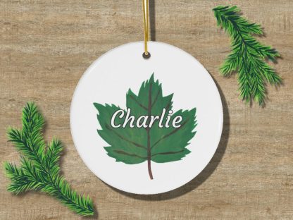 White, round ornament featuring a green leaf with "Charlie" in white script. Surrounded by pine branches on a wooden background. Festive and personalized.