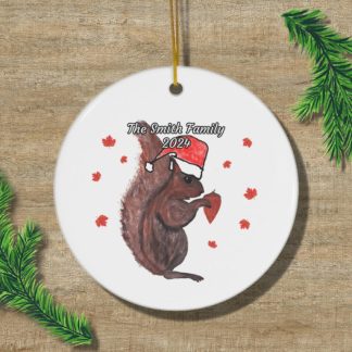 A round ornament on wood with a squirrel in a Santa hat holding a brown acorn, surrounded by small red leaves. Text reads "The Smith Family 2024." Pine branches frame the ornament, creating a festive holiday mood.