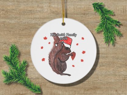 A round ornament on wood with a squirrel in a Santa hat holding a brown acorn, surrounded by small red leaves. Text reads "The Smith Family 2024." Pine branches frame the ornament, creating a festive holiday mood.