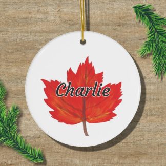 A round ornament featuring a red maple leaf with the name "Charlie" in black letters. It rests on a wooden surface, flanked by green pine branches.