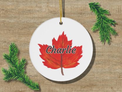 A round ornament featuring a red maple leaf with the name "Charlie" in black letters. It rests on a wooden surface, flanked by green pine branches.