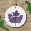 A white circular ornament featuring a purple leaf with the name "Sarah" in white script. Two green pine branches lie on a wooden surface nearby.