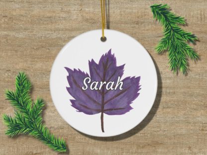 A white circular ornament featuring a purple leaf with the name "Sarah" in white script. Two green pine branches lie on a wooden surface nearby.