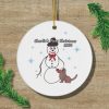 A white Christmas ornament with a snowman and dog illustration, adorned with snowflakes. Text reads "Charlie's First Christmas 2024." Festive greenery accents.