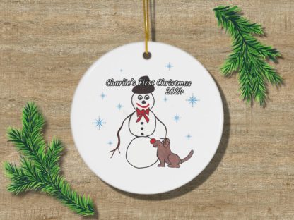 A white Christmas ornament with a snowman and dog illustration, adorned with snowflakes. Text reads "Charlie's First Christmas 2024." Festive greenery accents.