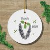 A white circular ornament with pine cones and holly design, featuring the name "Sarah" and the year "2024." Green pine branches surround it on a wooden background.