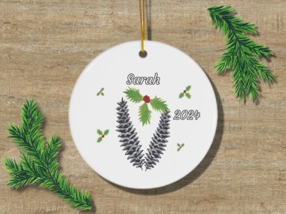 A white circular ornament with pine cones and holly design, featuring the name "Sarah" and the year "2024." Green pine branches surround it on a wooden background.