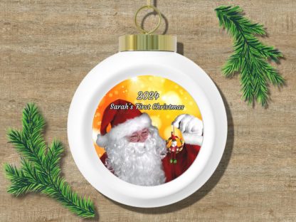 A white Christmas ornament on a wood texture background, featuring a smiling Santa holding a mini Santa ornament. Text reads "2024 Sarah's First Christmas." Pine branches accent the corners.
