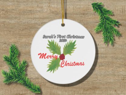 Round Christmas ornament on wood with "Sarah's First Christmas 2024" in black and "Merry Christmas" in red. Decorated with green holly leaves and red berries.