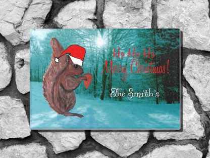 A festive holiday welcome mat features a squirrel in a red Santa hat standing on snowy ground against a blue-toned forest. Red text reads "Ho Ho Ho Merry Christmas!" and white text says "The Smith's."