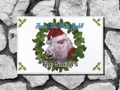 Holiday themed welcome mat with Santa Claus pointing, surrounded by a holly wreath. Text reads "Have You Been Good?" and "The Smith's." Festive and cheerful.