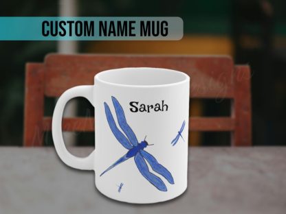 White mug featuring a large blue dragonfly and two smaller ones, with bold, artistic details. The personalized custom name "Sarah" is printed above in black letters, creating a personalized and whimsical design.