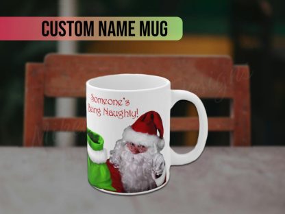 White mug featuring a Santa Claus graphic in a red suit, gesturing with his gloved finger. Text reads "Someone's Being Naughty!" in red, playful font.