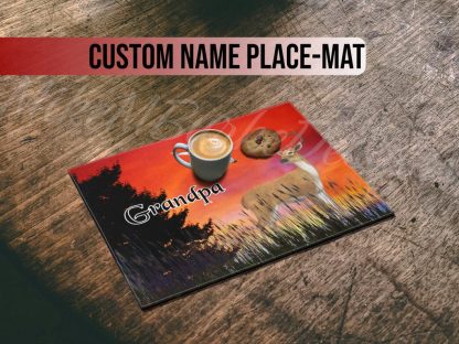 A placemat on a wooden table features a sunset scene with a deer and flying birds. The word "Grandpa" is written. A cappuccino and cookie rest on it.