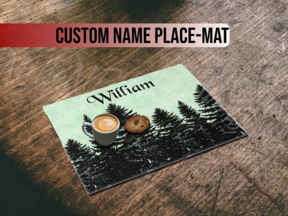 A coffee with latte art and a cookie sit on a placemat featuring pine trees and "William" in elegant script, resting on a rustic wooden table.