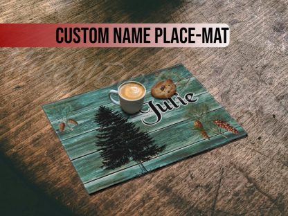 A rustic wooden table holds a teal placemat with pinecones, fir tree, and the name "Julie" printed. A latte and a chocolate chip cookie rest on top.