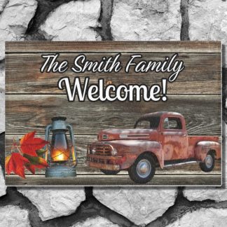 Welcome mat in a rustic wooden panel style on a gray stone step with "The Smith Family Welcome!" text. Features a vintage red truck, a glowing lantern, and autumn leaves, set against a rustic wooden panel design.