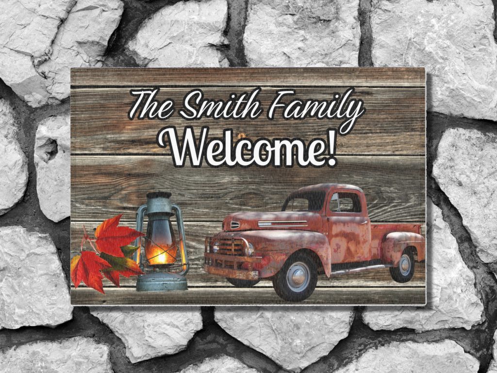 Welcome mat in a rustic wooden panel style on a gray stone step with "The Smith Family Welcome!" text. Features a vintage red truck, a glowing lantern, and autumn leaves, set against a rustic wooden panel design.