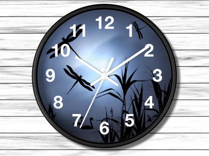 A wall clock with a black frame features silhouettes of dragonflies and reeds against a glowing blue background. White numbers and hands indicate the time.