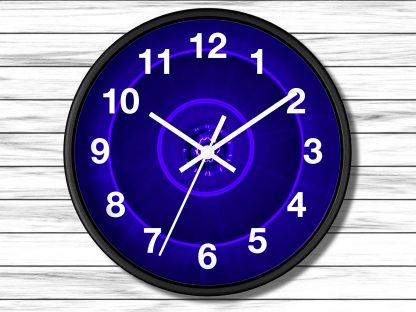 A wall clock with a black frame and glowing blue face, white numbers and hands. It hangs on a light wood-paneled wall. The design is modern and striking.