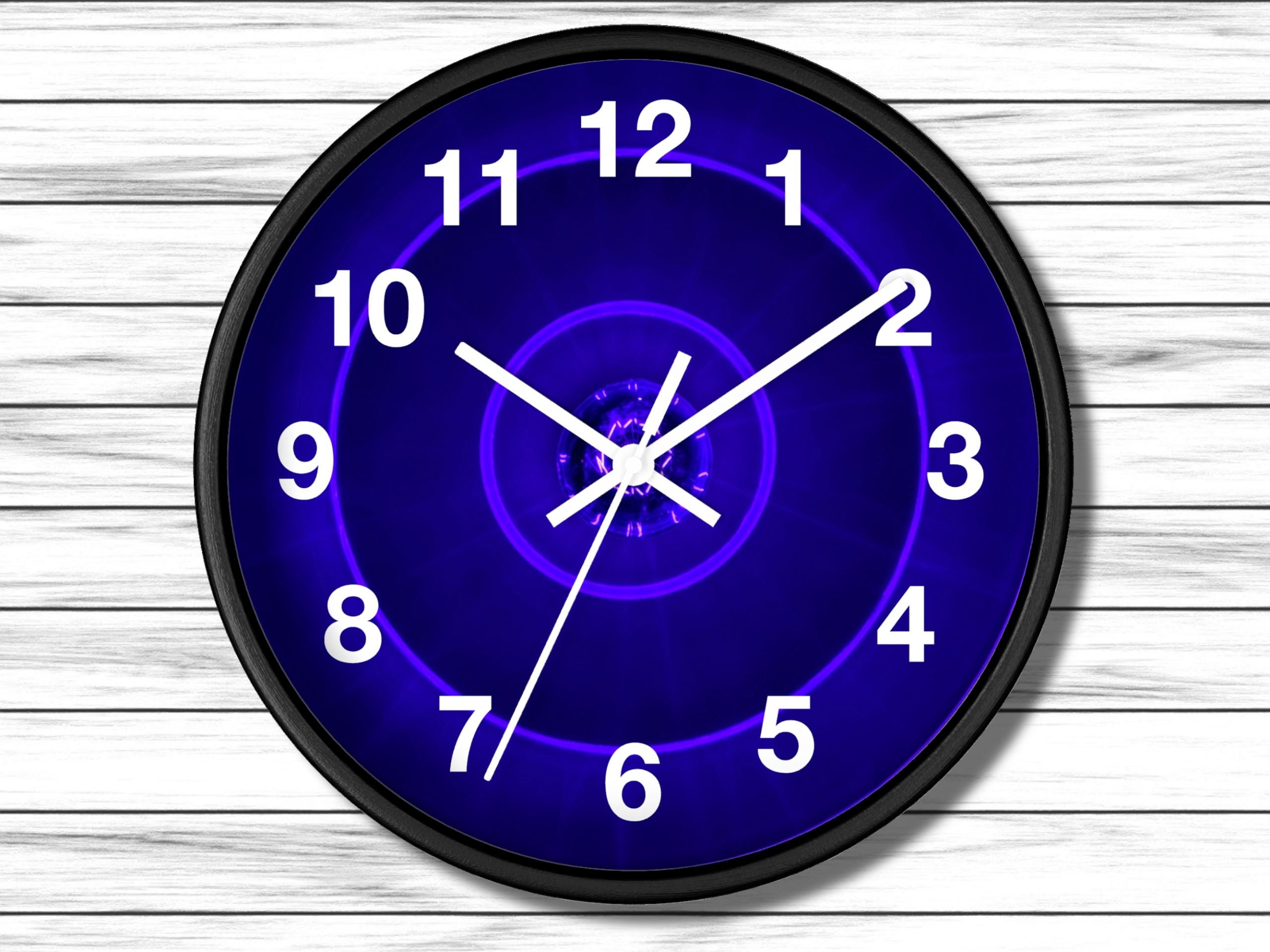A wall clock with a black frame and glowing blue face, white numbers and hands. It hangs on a light wood-paneled wall. The design is modern and striking.