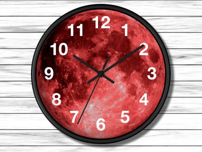 Round wall clock with a vibrant red moon face. Bold black hands point to 10:10. White numerals contrast against the vivid background. Wooden plank wall backdrop.