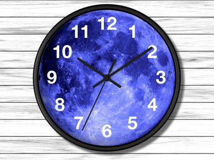 A round wall clock features a striking blue moon design, white numbers, and black hands, set against a wooden plank background, conveying a serene, cosmic vibe.