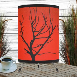 A rustic table lamp with a red shade featuring a black silhouette of a tree branch. It sits on a slatted wooden surface next to a white cup of coffee, with tall decorative grasses in the background.