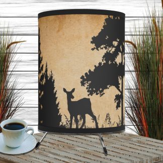 The image shows a wooden table with a cup of coffee and a decorative rustic lamp featuring a deer silhouette against a forest backdrop. Tall grasses are in the background.