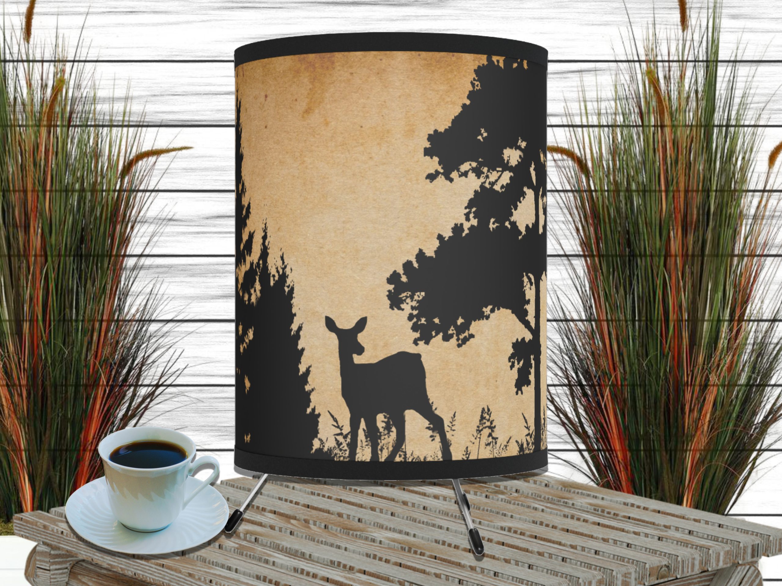 The image shows a wooden table with a cup of coffee and a decorative rustic lamp featuring a deer silhouette against a forest backdrop. Tall grasses are in the background.
