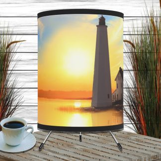 Table with a decorative nautical lighthouse-themed lamp displaying a serene sunset over water. A cup of coffee is on the table, framed by tall grasses.