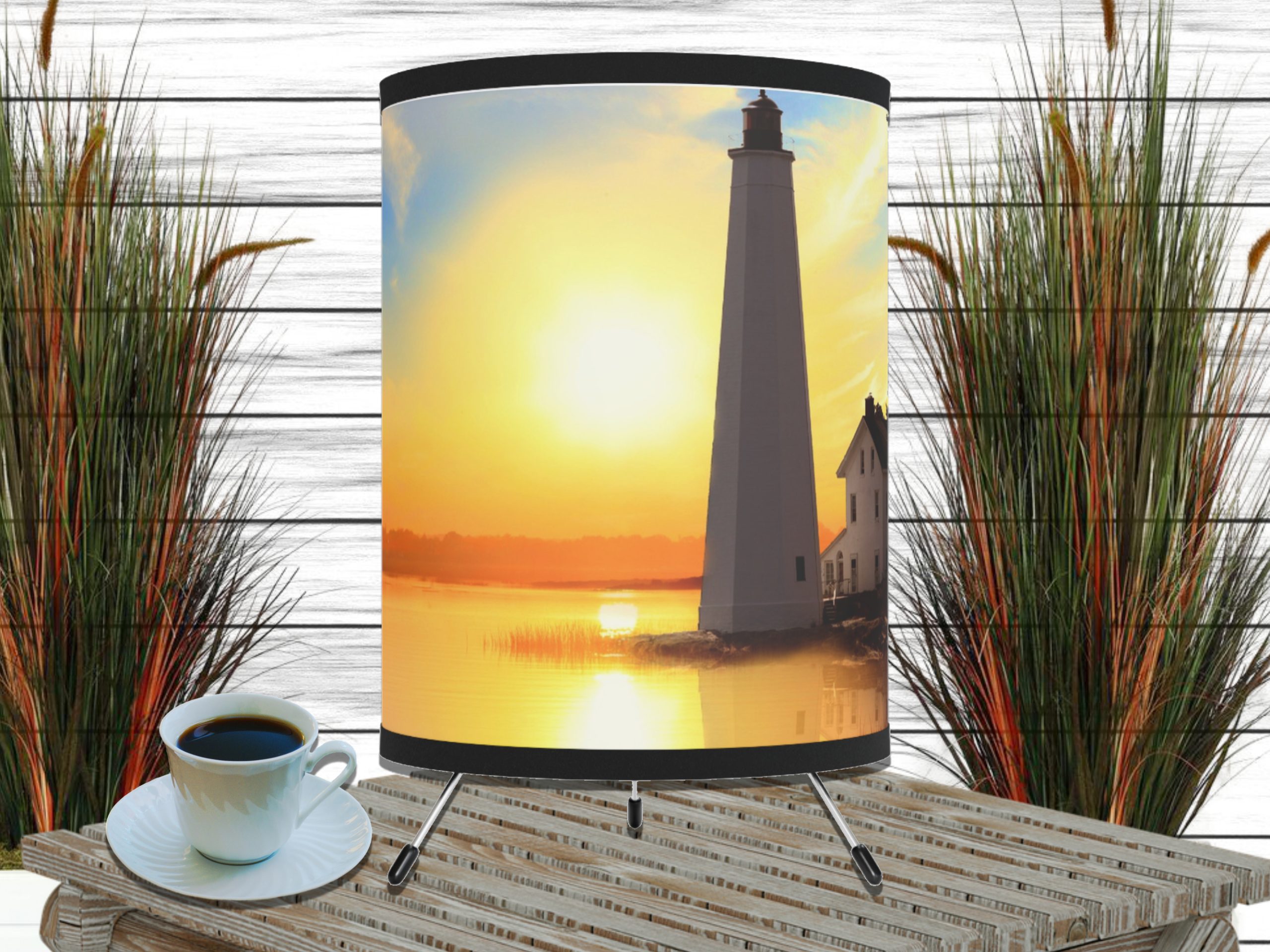Table with a decorative nautical lighthouse-themed lamp displaying a serene sunset over water. A cup of coffee is on the table, framed by tall grasses.