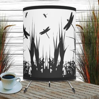 Silhouette-themed table lamp with dragonfly and grass designs, set on a wooden surface next to a cup of coffee. Tall grass decor in background. Calm ambiance.