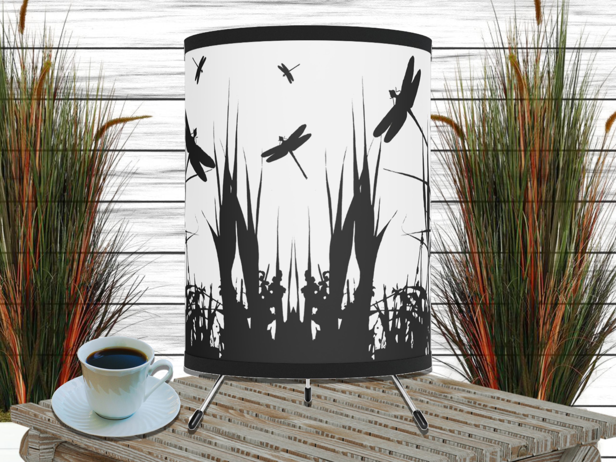Silhouette-themed table lamp with dragonfly and grass designs, set on a wooden surface next to a cup of coffee. Tall grass decor in background. Calm ambiance.