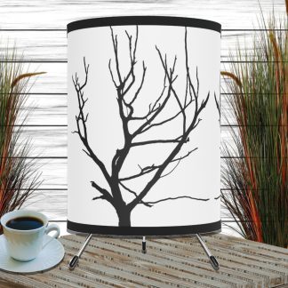 A rustic table lamp with a black tree branch silhouette on a white shade stands on a wooden surface. Beside it is a white cup of black coffee. In the background, tall grass with green and orange tones contrasts against white wooden panels, creating an earthy, tranquil ambiance.