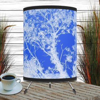 A stylish lamp with a white tree silhouette against a vivid blue background stands on a striped wooden table. Next to it, a cup of black coffee and tall grass plants in soft, natural light.